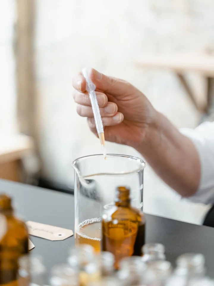 Close-up of a hand holding a dropper over a beaker in a lab, with amber bottles and glassware in the background.