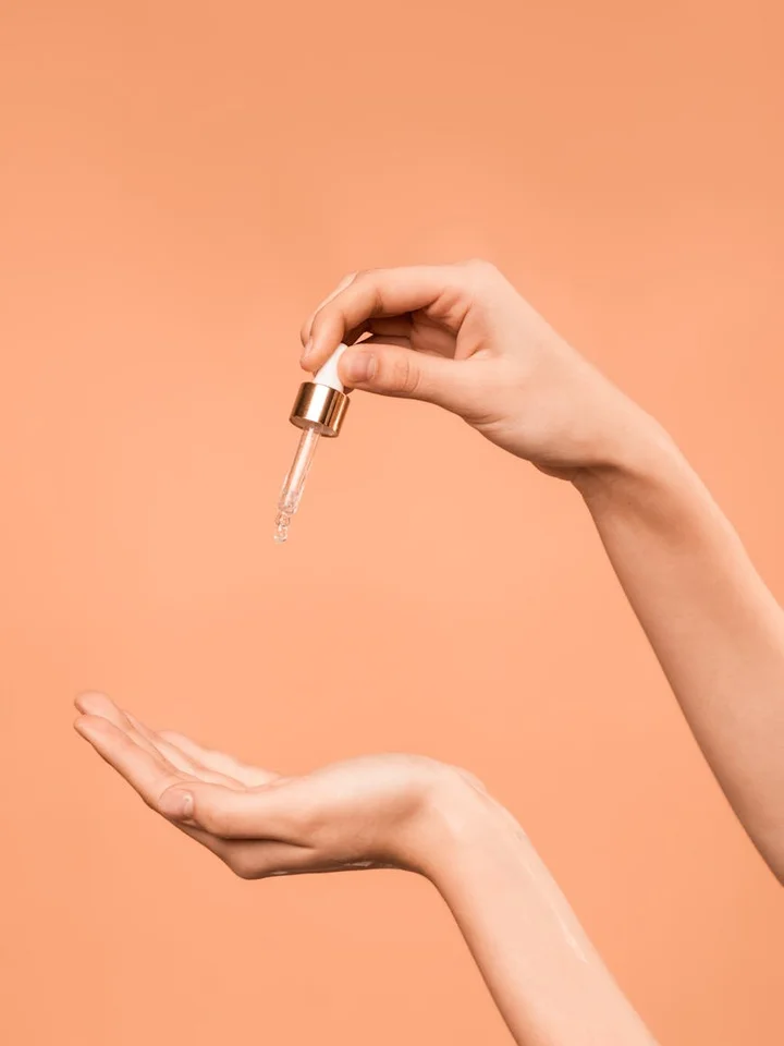 Close-up of a hand holding a dropper above an open palm, about to dispense retinol onto the skin against a peach background