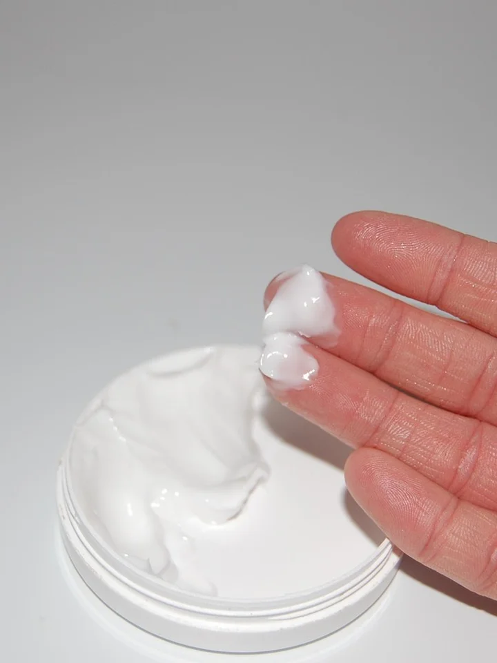 Close-up of a hand with a dollop of white cream on the fingertips above an open jar of moisturizer