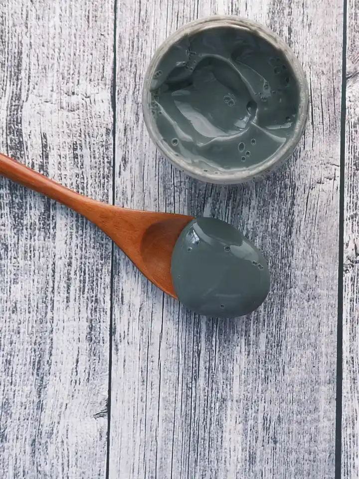 A small jar of gray cream with a wooden spoon on a light wood surface, representing topical retinoids for skincare.