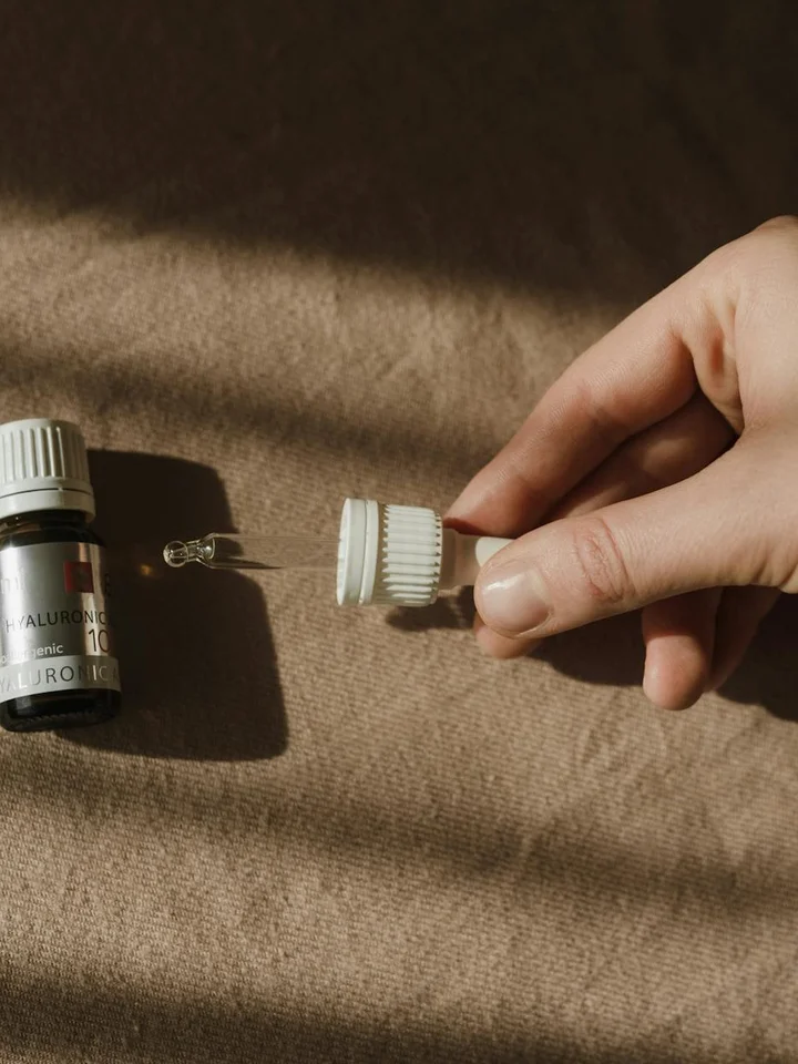 Close-up of a hand holding a small cap and dropper from a salicylic acid bottle on a brown fabric surface.