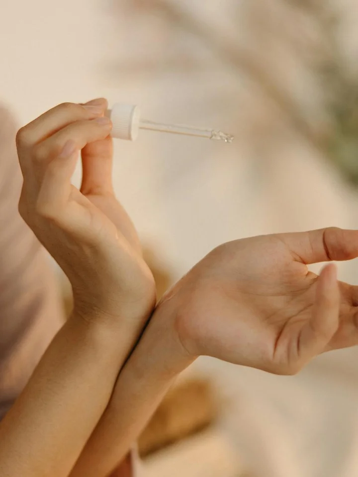 Close-up of hands with a dropper bottle dispensing serum onto the forearm during a skincare routine.