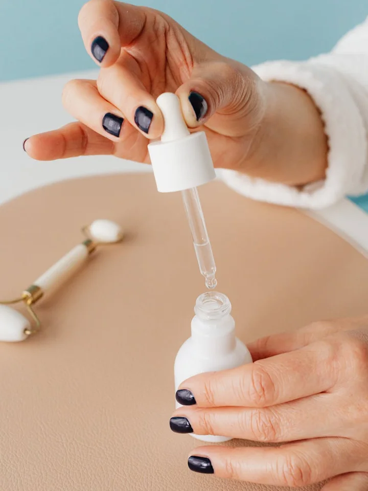 A skincare setup showing a hand with dark nail polish holding a dropper and dispensing serum onto the skin.