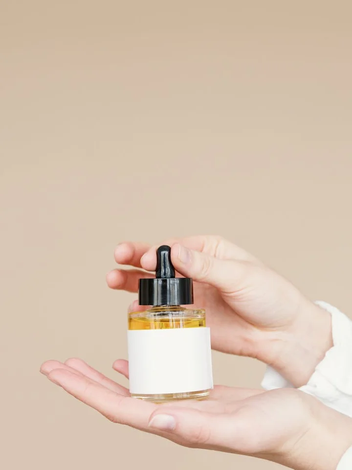 A small glass dropper bottle held between two hands, set against a neutral beige background.