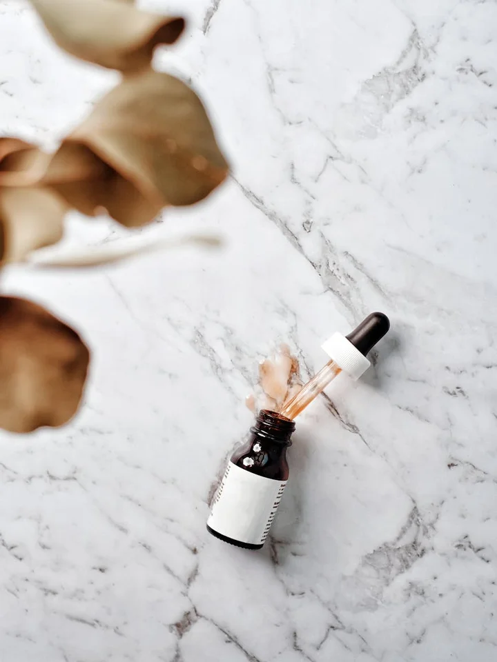 Dropper bottle on a white marble surface with dried leaves in the foreground, symbolizing skincare preservatives and natural derivatives.