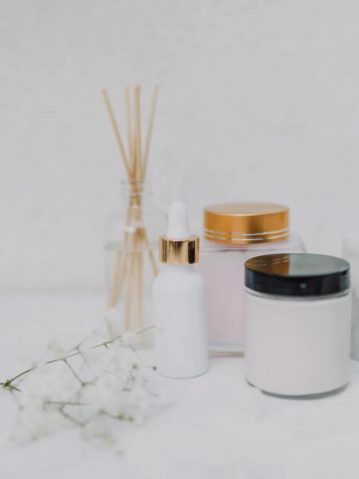 Minimal skincare setup featuring a white dropper bottle and two jars on a pale surface, with diffuser sticks and white flowers in the foreground.