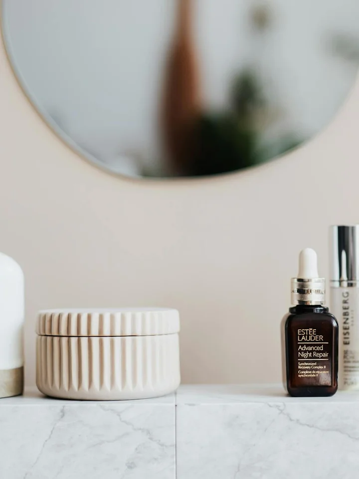 A marble countertop with skincare products: a ribbed white jar and a dark dropper bottle, with a blurred mirror in the background.