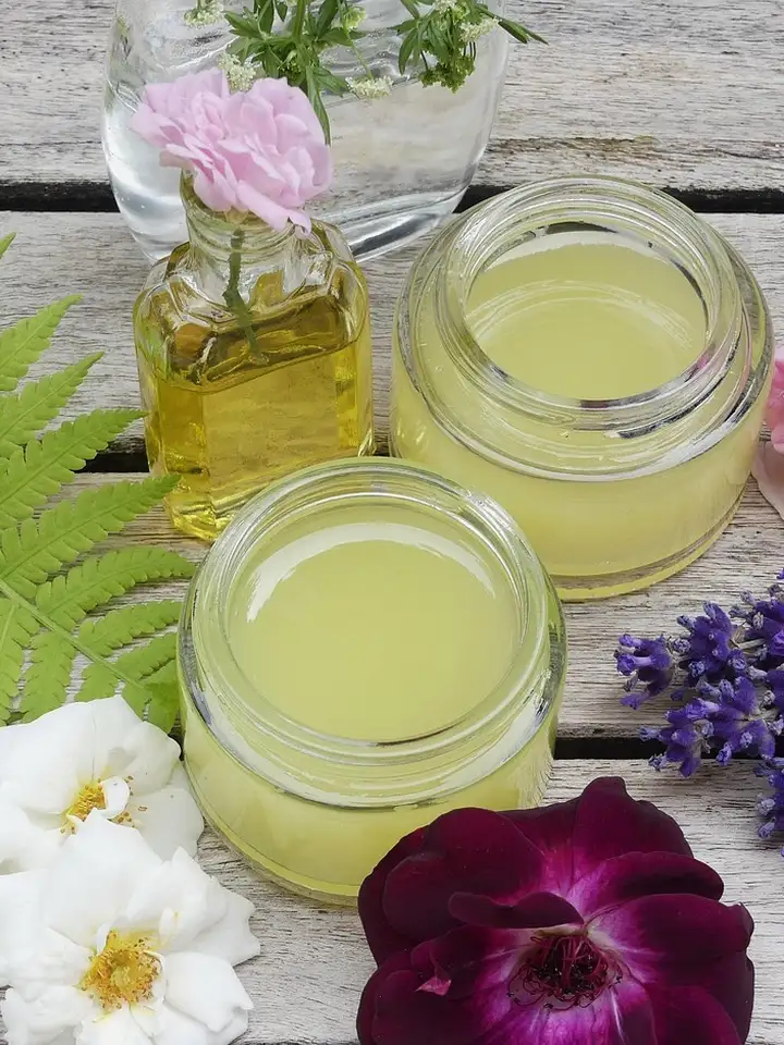 Three glass jars of pale yellow skincare oil and butter on a wooden surface, with pink and white flowers and green ferns nearby.