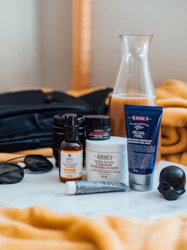 An assortment of skincare products on a marble surface, including a blue Kiehl's tube, a white jar, a small amber bottle with a dropper, and sunglasses, with a soft orange towel in the foreground.