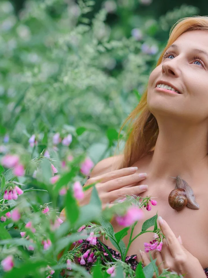 A woman in a garden with a snail on her shoulder, looking up with a gentle smile.