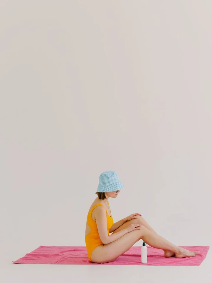 A person in a yellow swimsuit and light blue knit hat sits cross-legged on a pink towel with a sunscreen bottle nearby.