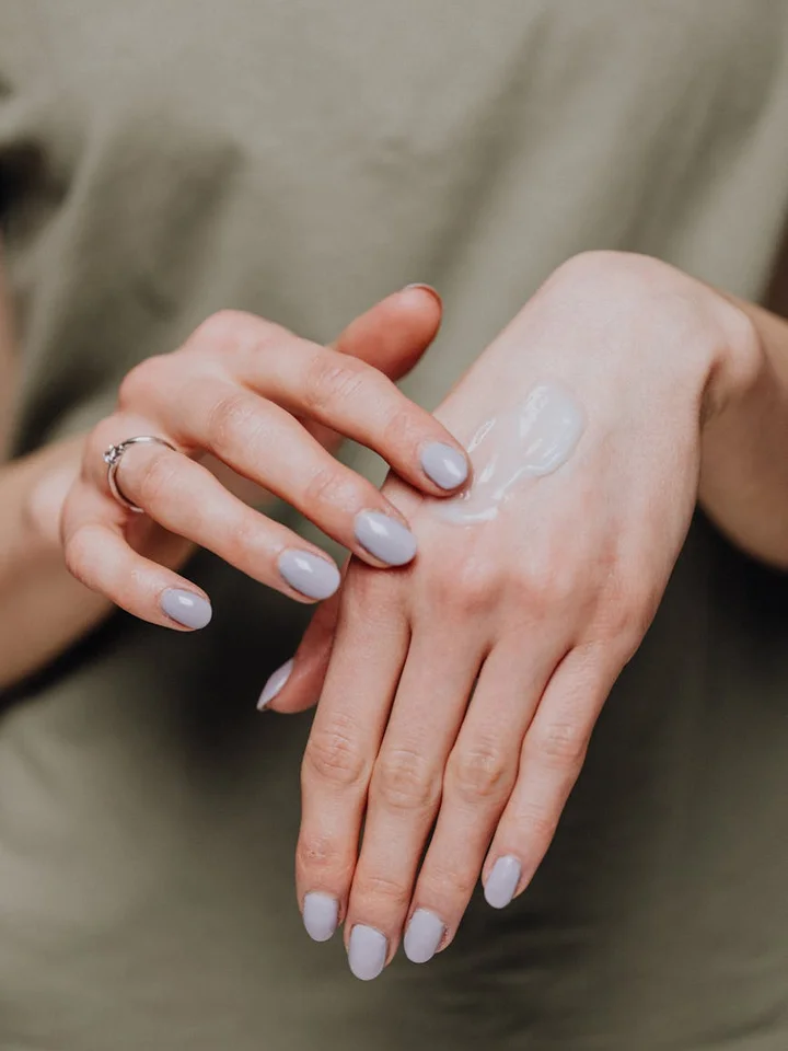 Close-up of hands with a small dollop of skincare product on the back of one hand; nails painted in pale lavender.