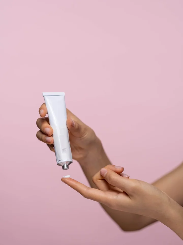 Close-up of hands dispensing a small amount of cream from a tube onto a fingertip against a pink background