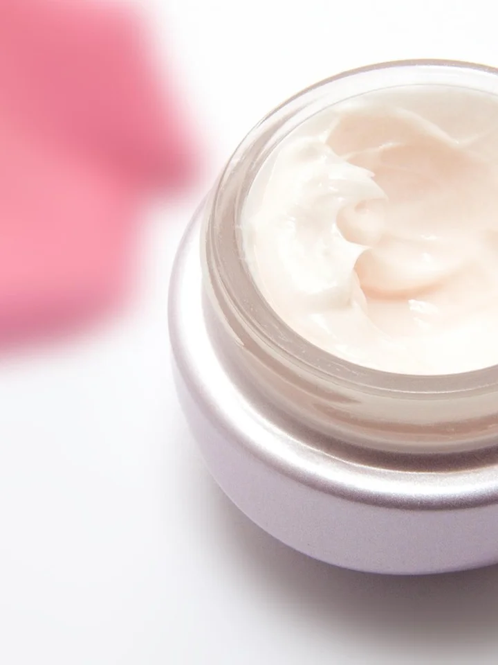 A small jar of pale cream with a pink blurred background on a white surface.