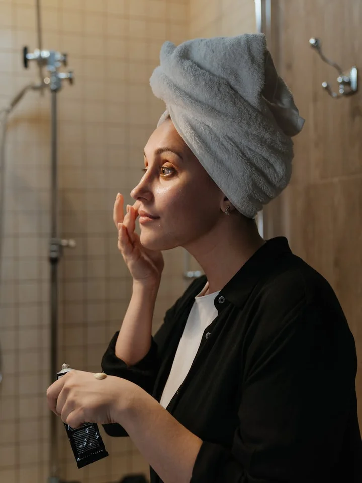 Person with a towel wrapped around their head applying skincare product in a bathroom