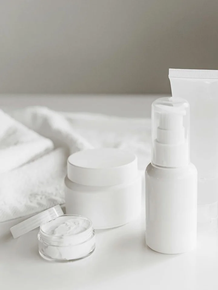 Assorted white skincare products, including a pump bottle and small jars, arranged on a white surface.