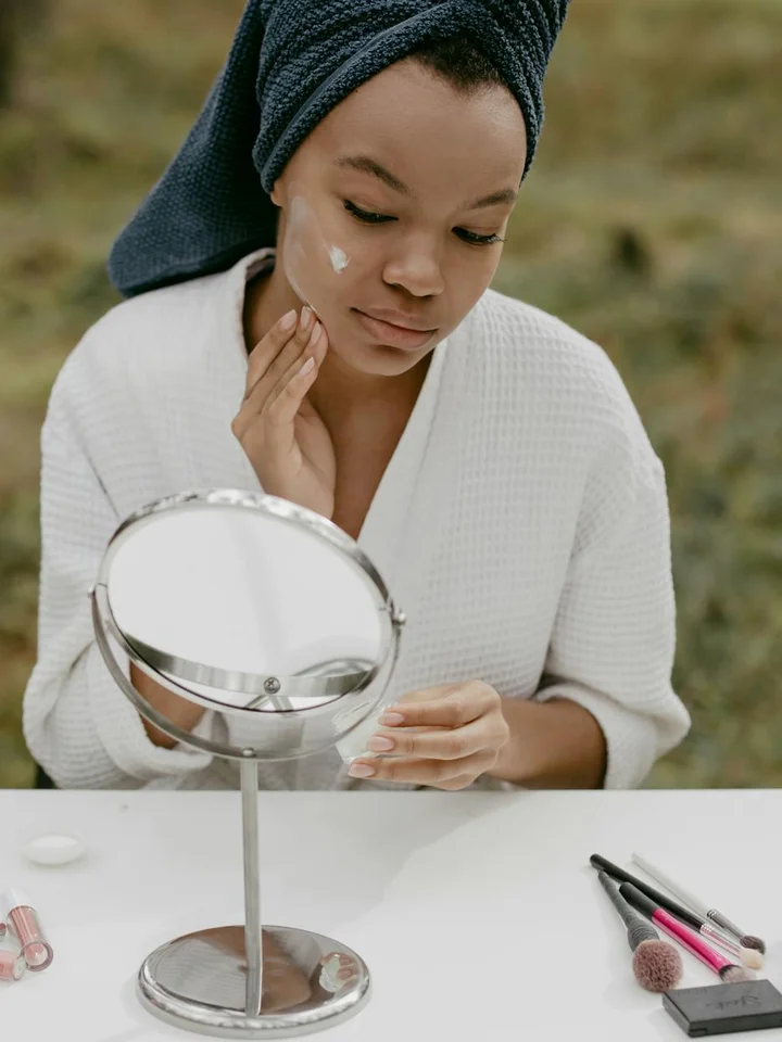 Woman wearing a white robe with a towel wrapped around her head applies a small amount of cream to her face while looking into a handheld mirror outdoors.