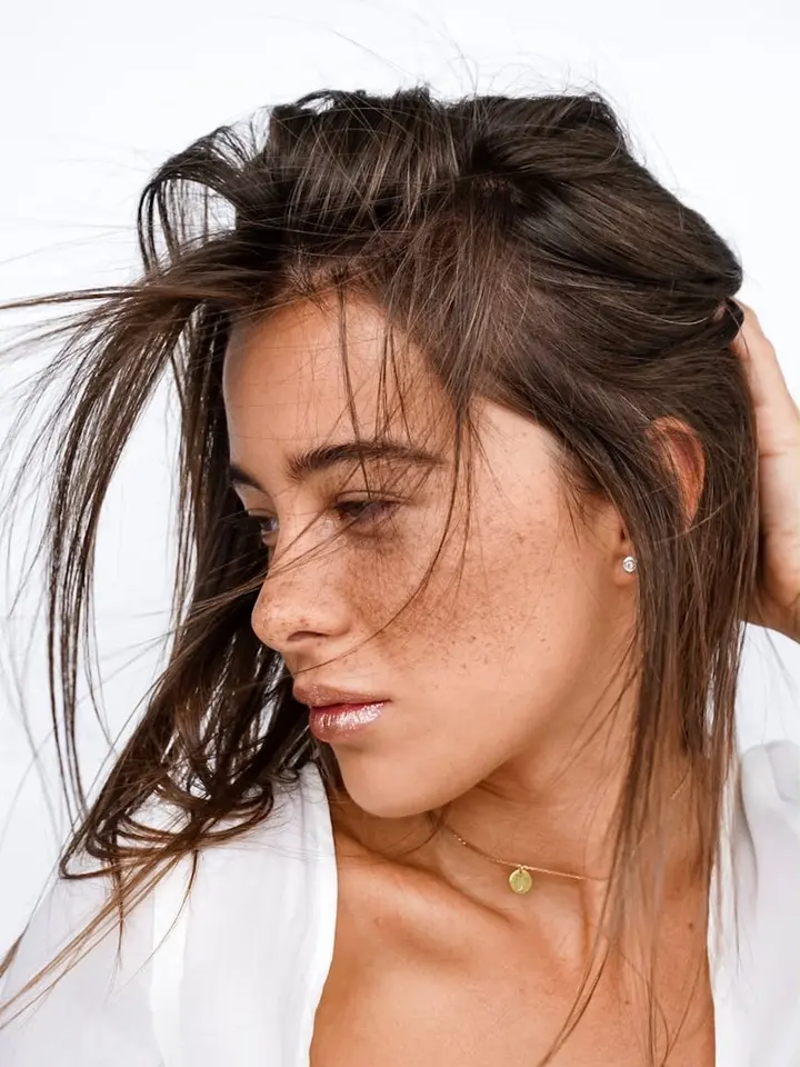 Side-profile close-up of a freckled woman with brown hair, wearing a white top.