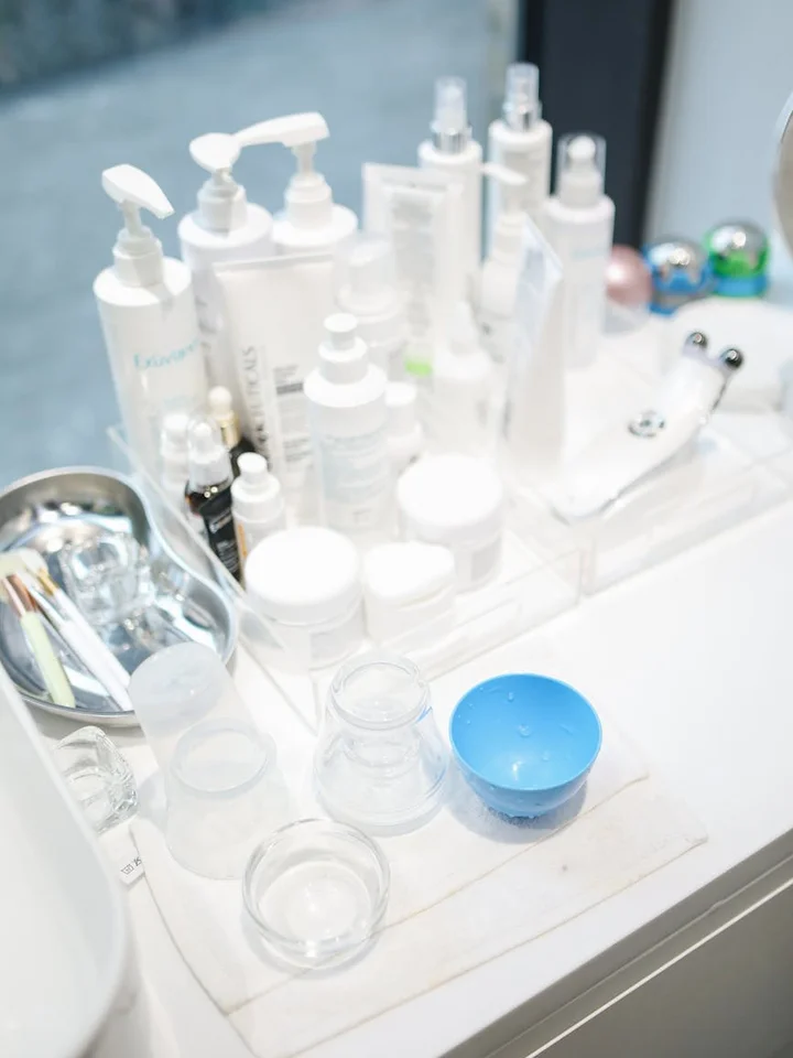 Assorted skincare bottles and jars arranged on a white bathroom vanity, with a blue bowl in the foreground.