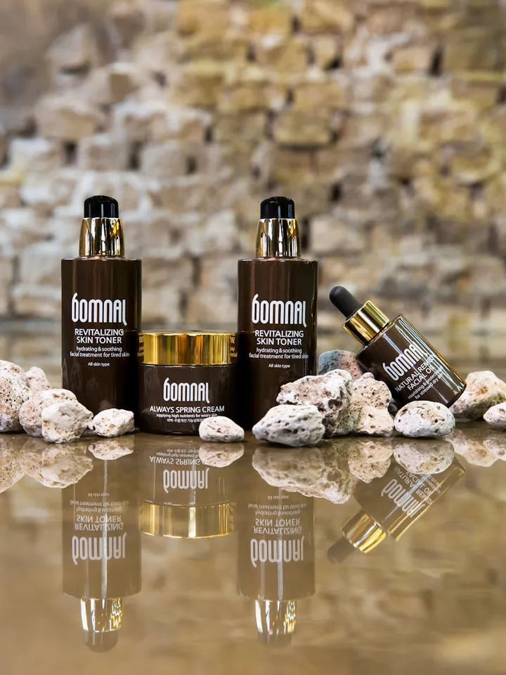 Row of dark skincare bottles with gold caps on a rocky surface by water, with reflections on the surface.