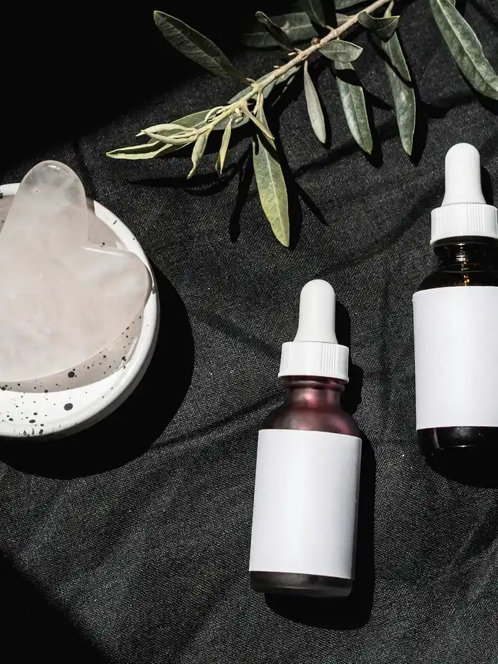 Dark fabric background with two dropper bottles of serums and a heart-shaped stone on a dish, with an olive branch nearby.