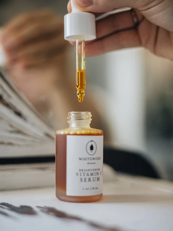 Close-up of a dropper releasing orange vitamin C serum into a small amber bottle labeled Vitamin C Serum, with a blurred skincare setup in the background.