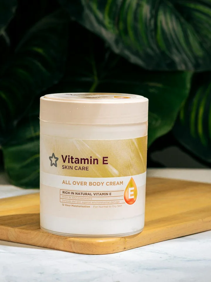 A jar labeled Vitamin E Skin Care All Over Body Cream sits on a wooden board with green tropical plants in the background.