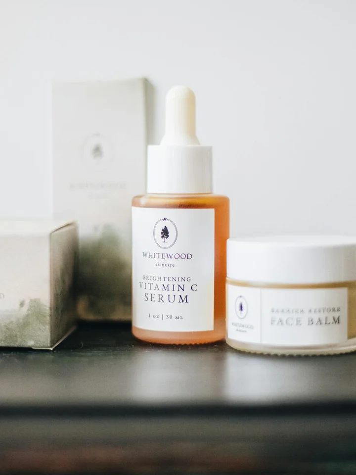 Dropper bottle labeled Vitamin C Serum and a small jar of cream on a dark surface, illustrating skincare products for layering.