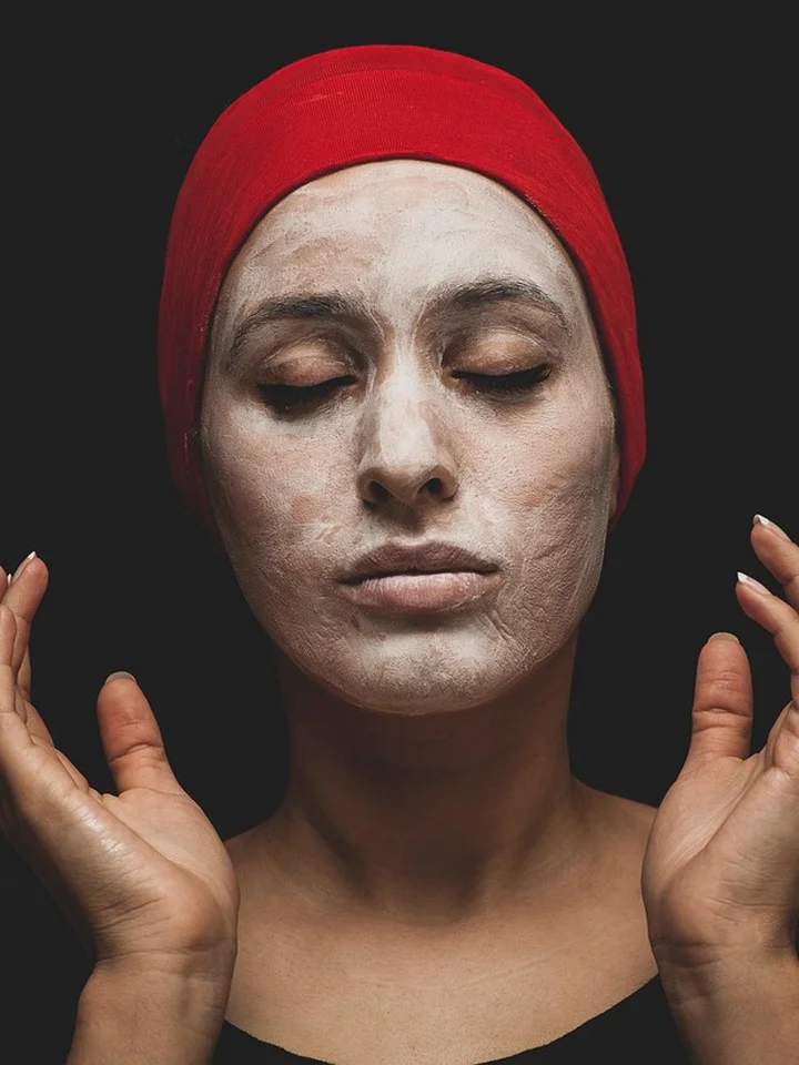 Woman with a white facial mask and red head wrap, eyes closed, practicing skincare.