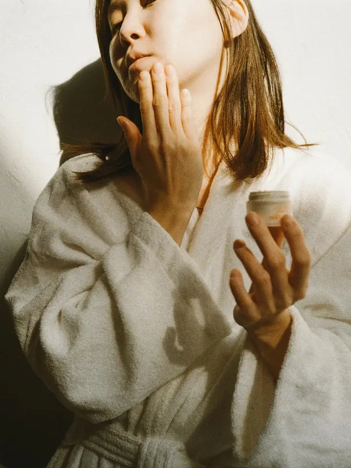 Person in a white bathrobe holding a small skincare jar and touching their lips with their other hand.