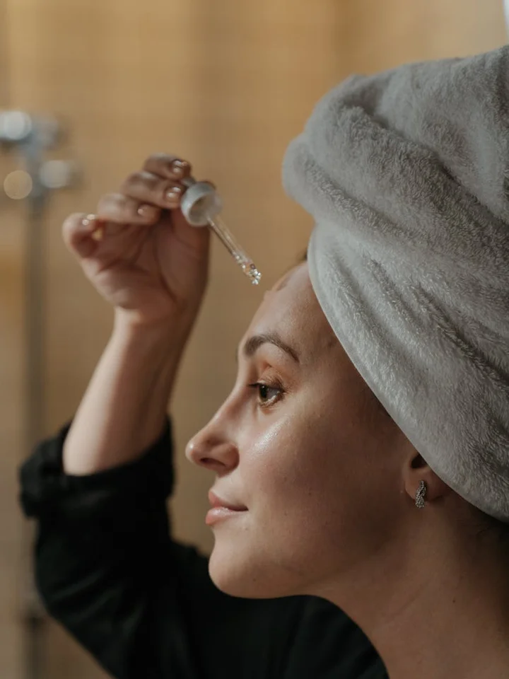 Woman with a towel wrapped around her head applying skincare serum with a dropper to her face.