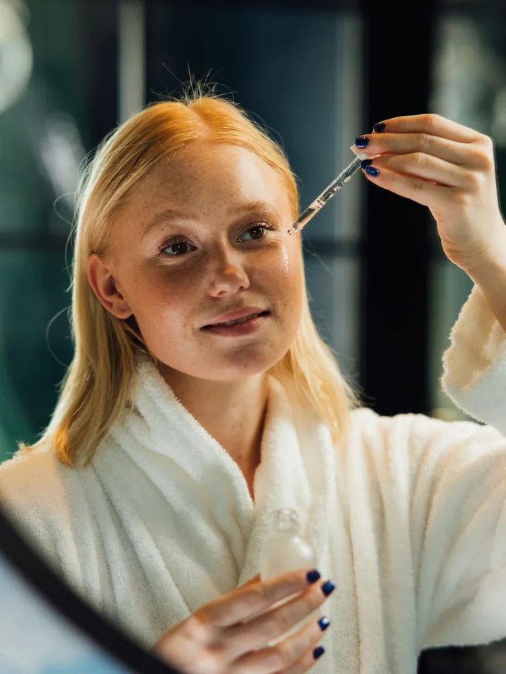 Person in a white bathrobe holding a skincare dropper and bottle, preparing to apply serum.