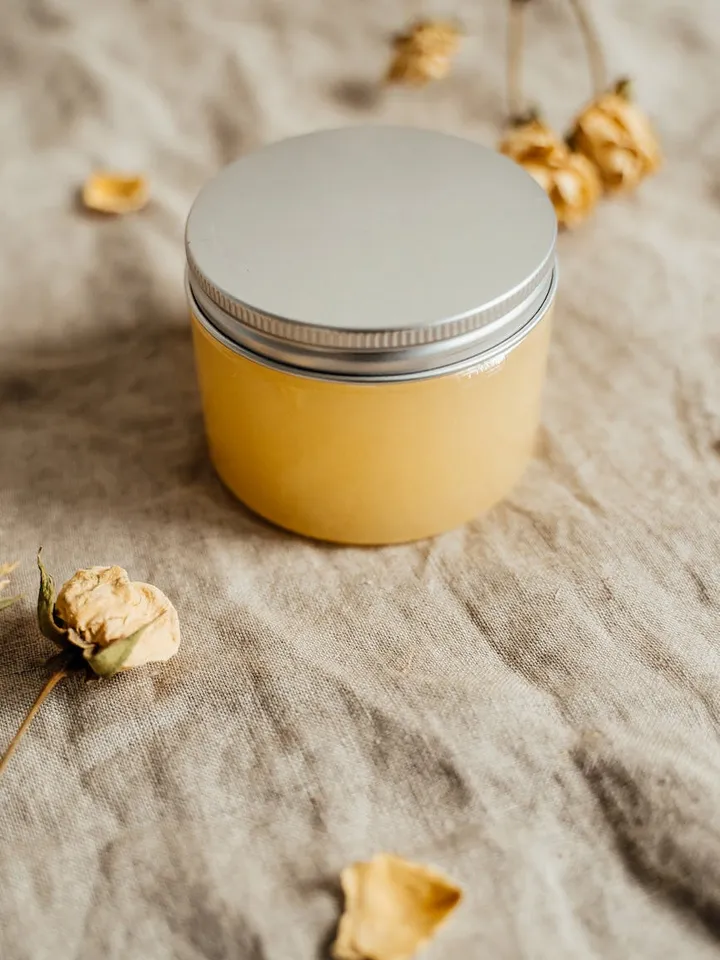 Small jar of yellow topical cream with a silver lid sits on beige fabric, with dried flowers around it.
