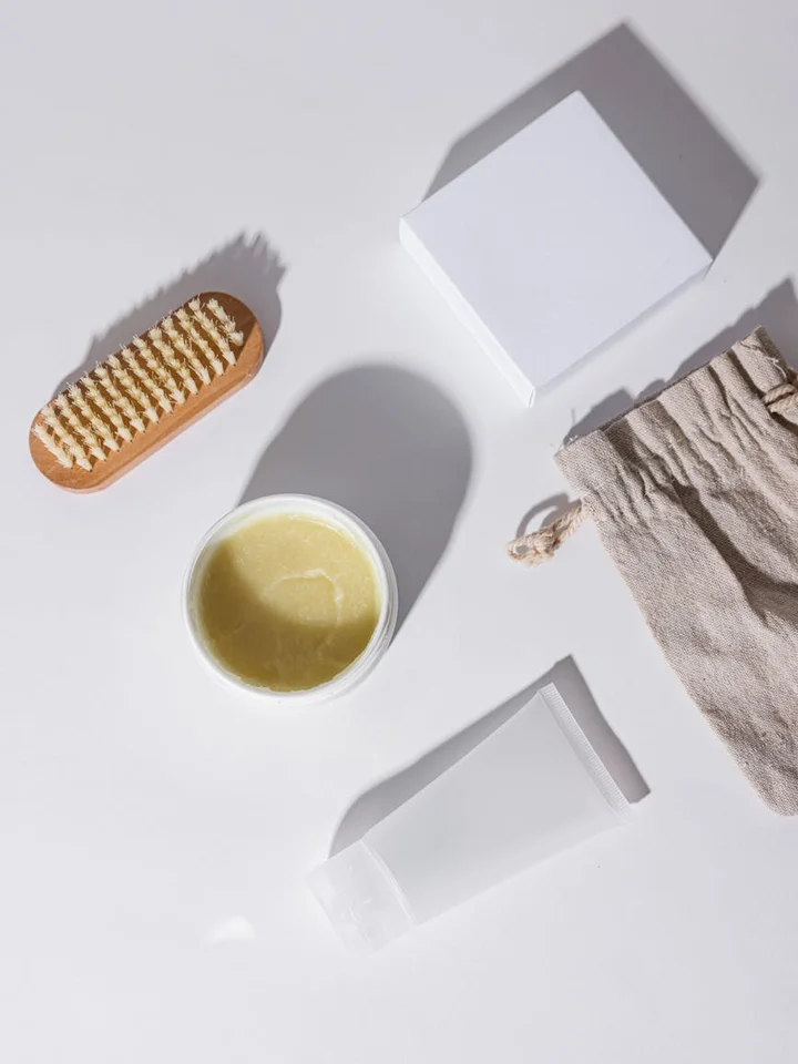 Flat lay of skincare items including a brush, a jar of cream, two white tubes, a cloth, and a box on a white surface.