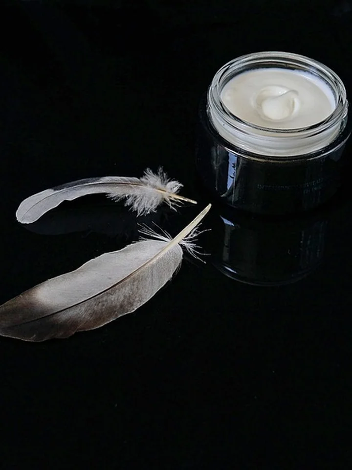 A small glass jar of cream on a dark surface with two white feathers beside it, suggesting gentle skincare and caution as you begin using adapalene.