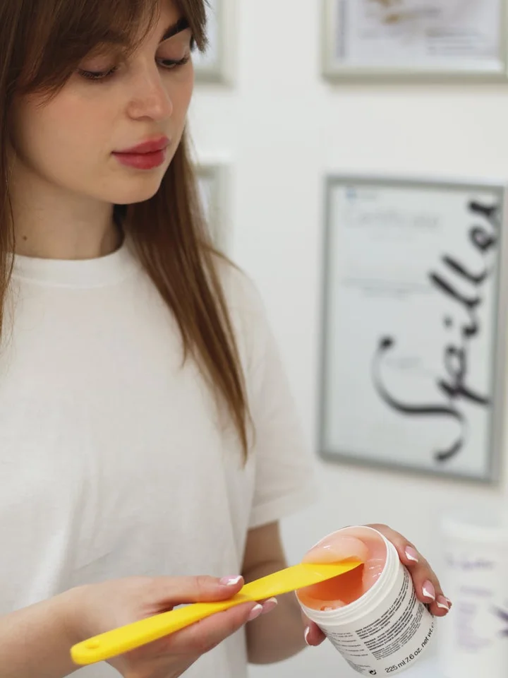 Person in a white shirt holds a jar of skincare product with a yellow spatula, preparing to apply it.