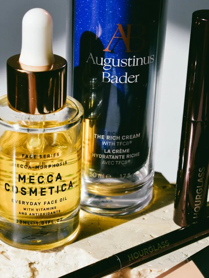 Close-up of skincare products on a surface: a Mecca Cosmetica Everyday Face Oil dropper bottle, an Augustinus Bader The Rich Cream bottle, and an Hourglass cosmetic pencil.