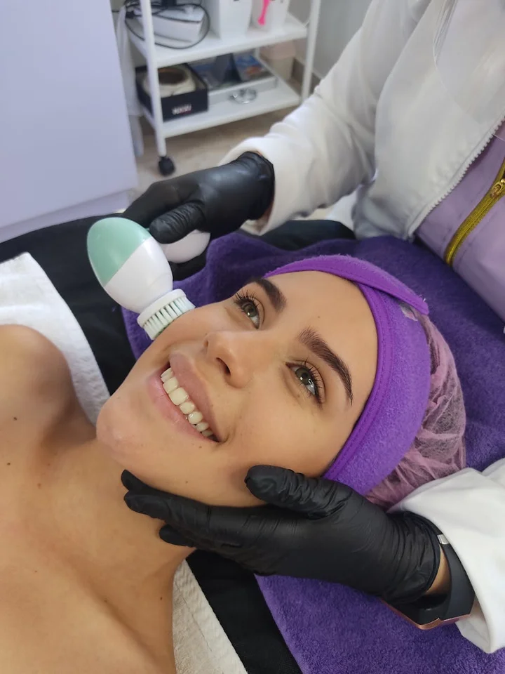 A clinician wearing black gloves uses a handheld facial device on a smiling patient with a purple headband, lying on a treatment bed