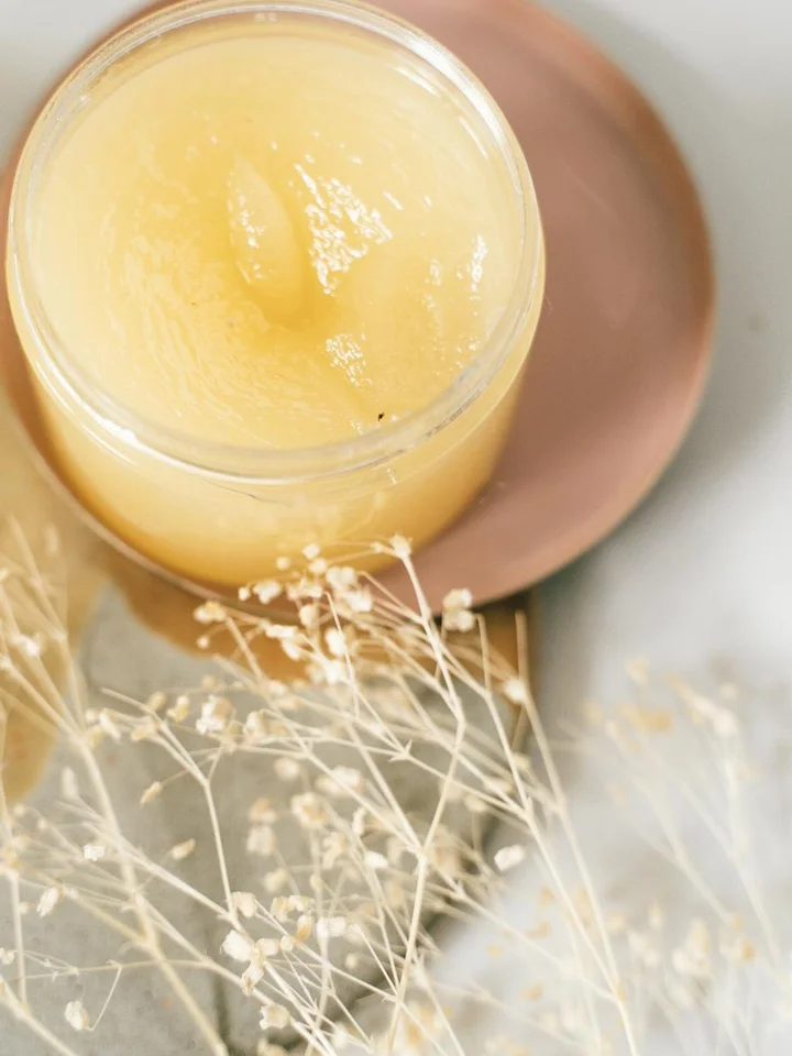 Open jar of pale yellow adapalene cream on a soft neutral surface with delicate dried flowers nearby.