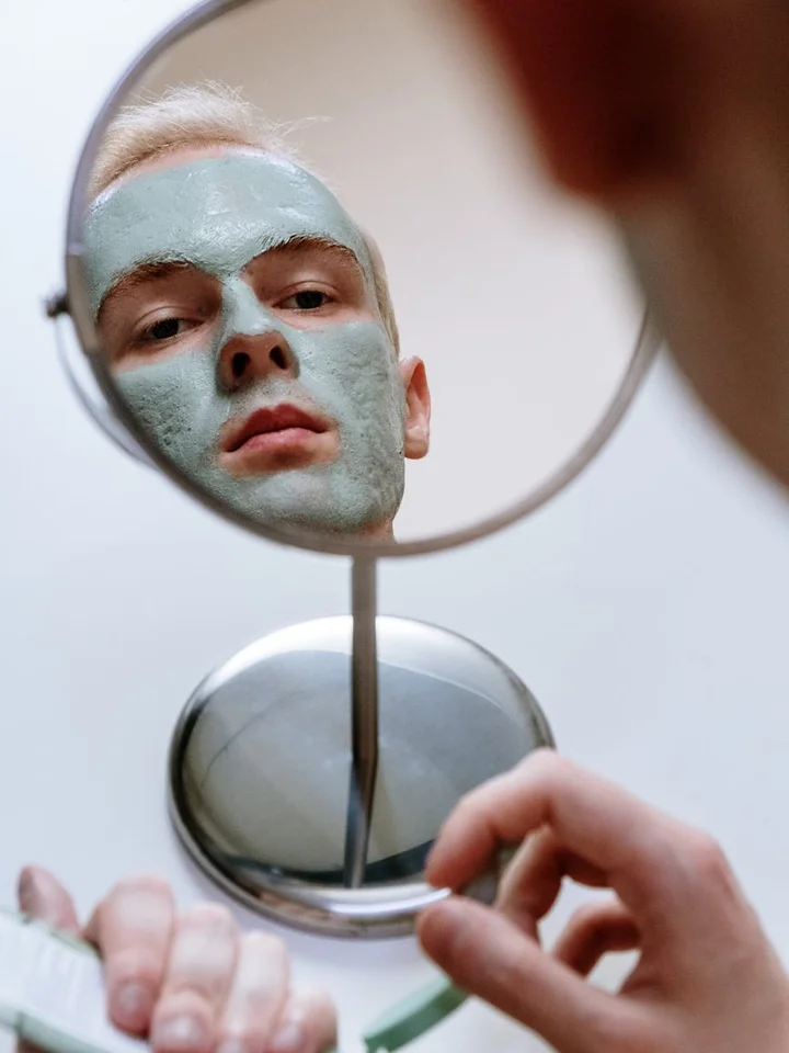 Person wearing a green facial mask looking at their reflection in a round handheld mirror during a skincare routine.