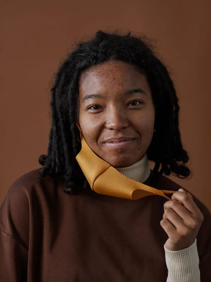Portrait of a young person with dreadlocks, wearing a brown sweater and a yellow inner collar, smiling, against a warm brown background, adjusting the collar.