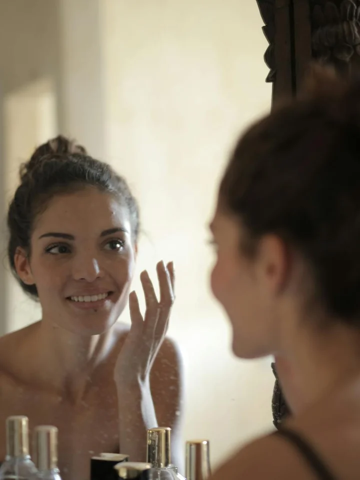 Woman smiling while looking at her reflection in a bathroom mirror as she applies skincare.