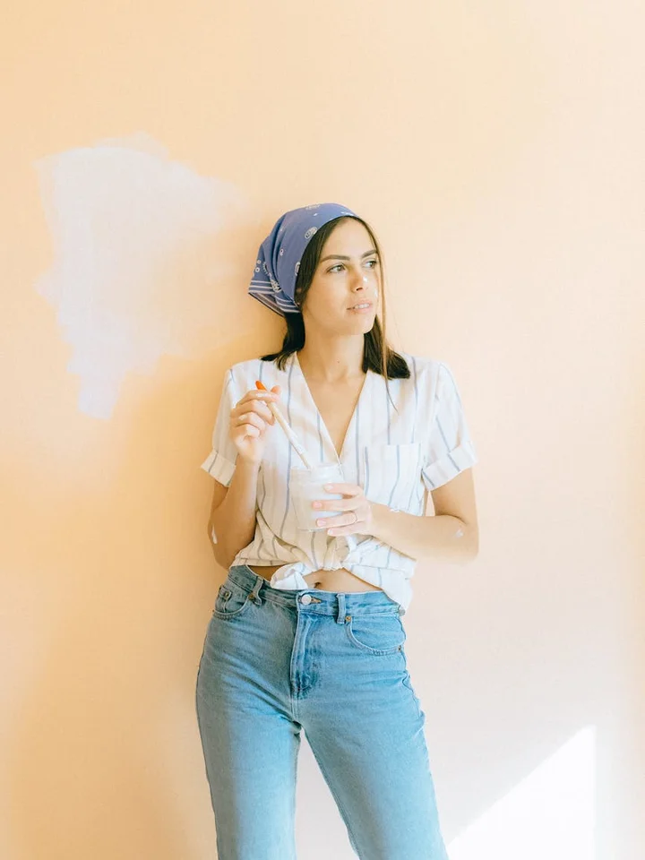A woman stands against a peach-colored wall, wearing a light striped shirt and a blue headscarf, looking off to the side.