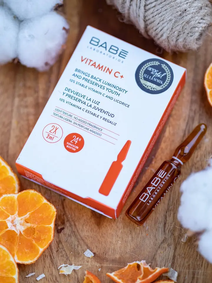 A BABE Vitamin C serum box and a small amber glass dropper bottle sit on a warm wood surface, surrounded by orange slices, cotton balls, and a beige fabric.