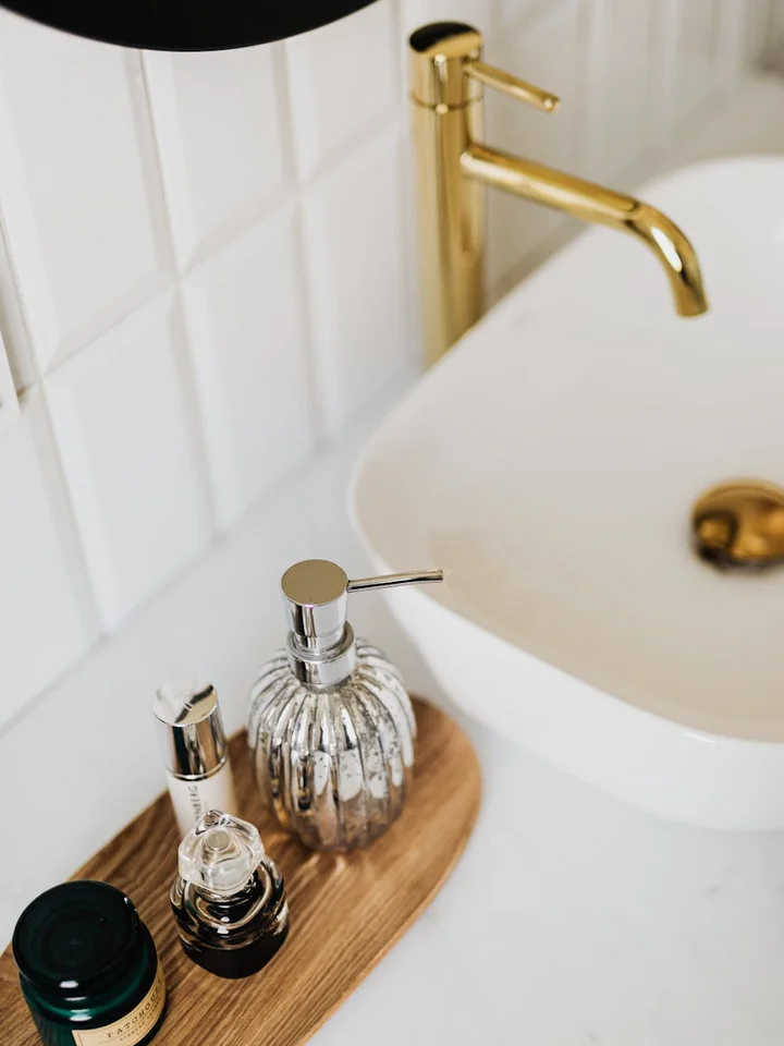 Bathroom countertop with a white sink and gold faucet, displaying a wooden tray with assorted skincare bottles and containers.