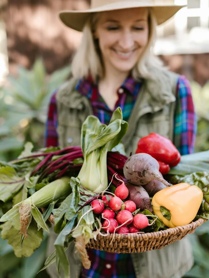 A smiling woman wearing a hat holds a basket of fresh vegetables, including beets, radishes, and a yellow bell pepper.
