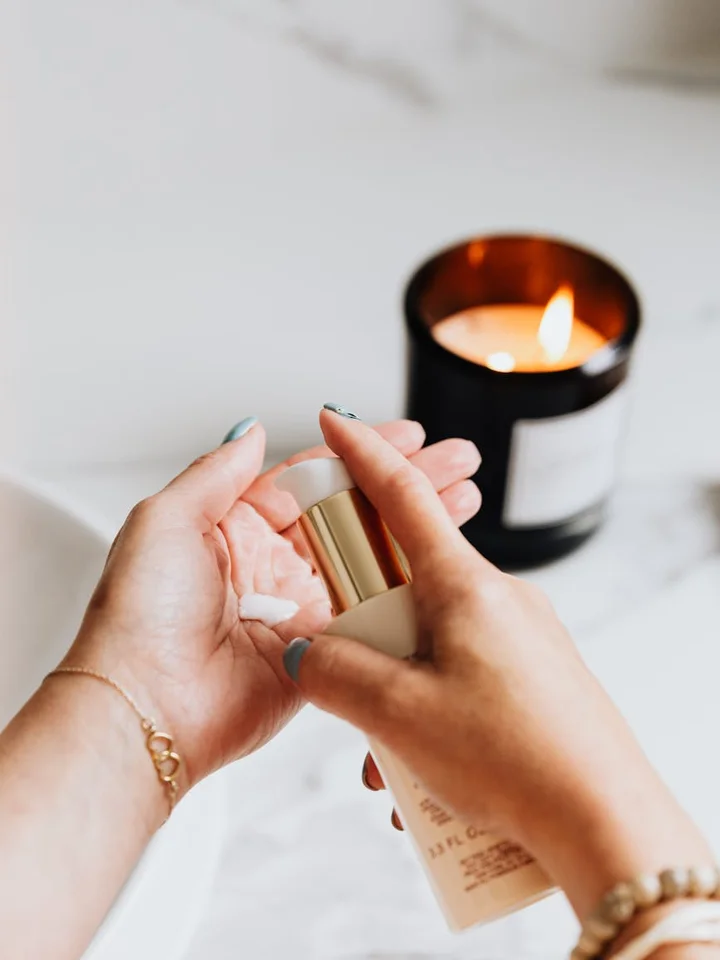 Close-up of hands holding a skincare pump bottle in a bright bathroom with a candle burning in the background.