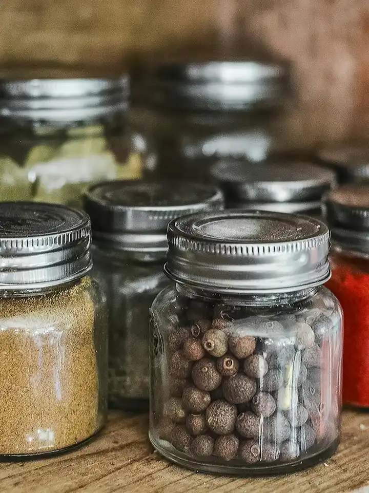 Assorted glass jars with metal lids containing ground powders and seeds on a wooden surface.
