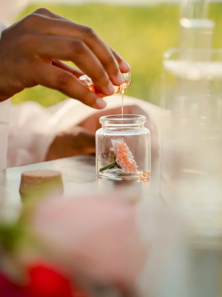 Close-up of hands using a dropper to add oil from a vial into a small glass jar with a pinkish flower inside, blurred lab-like surroundings and natural light.