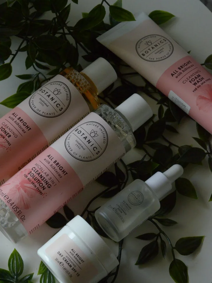 Botanics All Bright skincare bottles arranged on green leaves, featuring pastel pink packaging and a dropper bottle